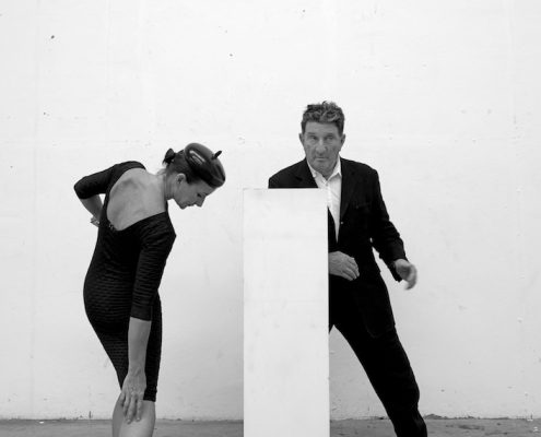 Black and white photo of artist Bruce McLean and his daughter designer Flora McLean dancing around an empty white plinth